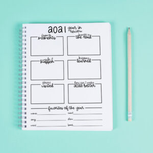 This image represents the free printable that you can download in this blog post. There is a notebook on a teal blue background with a white pencil next to it. The notebook is open to the free printable available in this blog post - the free 2021 year in review printable.
