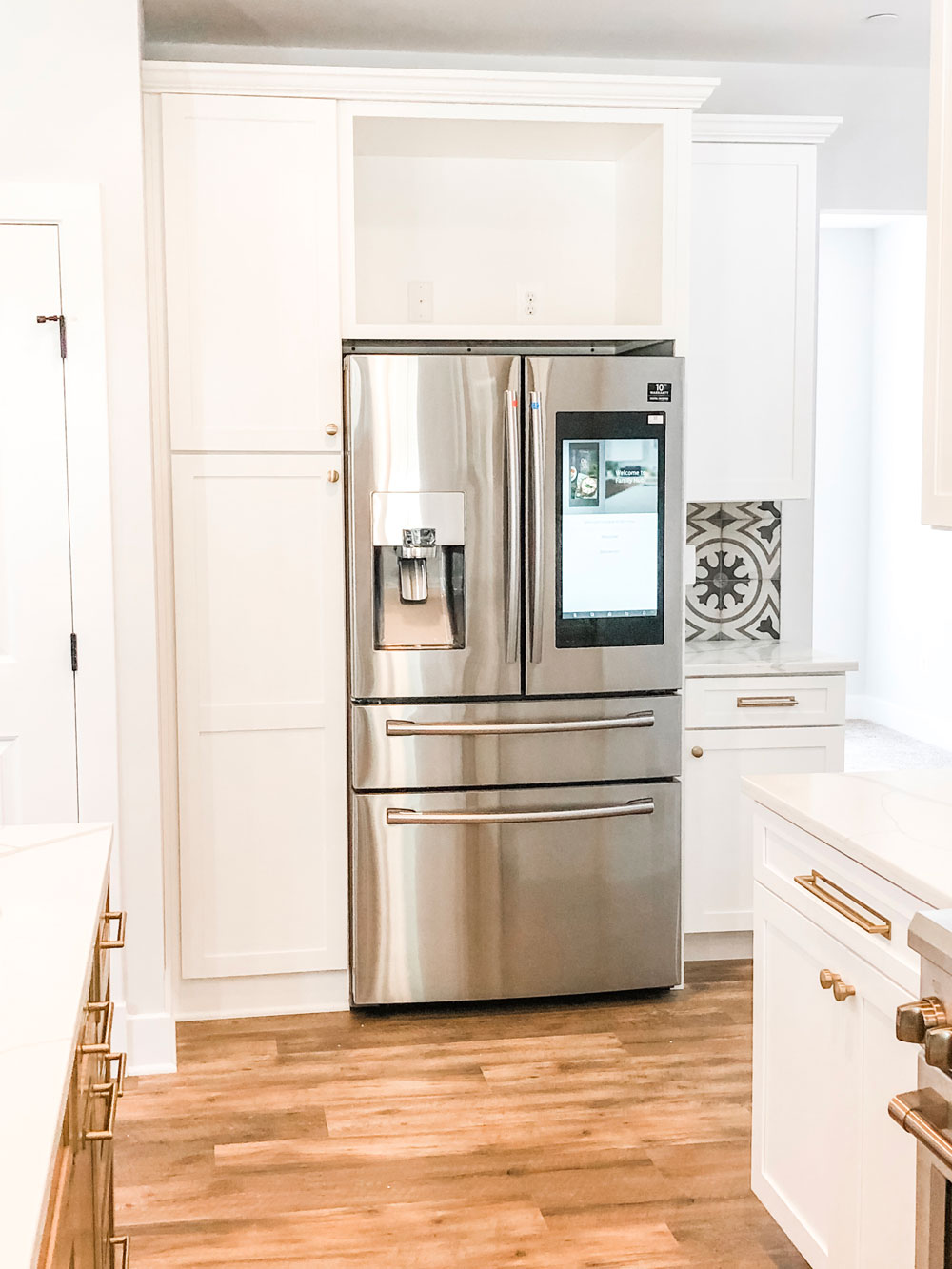 Picture from a custom home tour of a kitchen. This picture shows a refrigerator surrounded by white cabinetry. The fridge is a smart fridge with a screen like a tablet or phone.