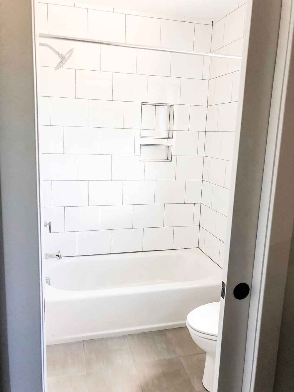 Photo from a custom home tour. This photo shows a picture of a bathtub with large white tile laid in a subway pattern with dark gray grout. 