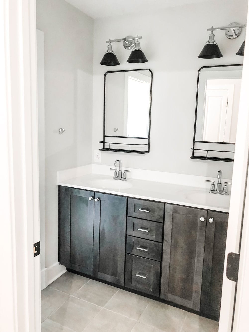 Custom home tour photos. This photo shows a double bath vanity. The bath cabinet is a dark wood grain and it has a solid white marble top. There are two black mirrors - one above each sink. There is also a light above each sink that is a two light fixture with black shades and chrome for the rest of the fixture. There are chrome handles and chrome faucets. 