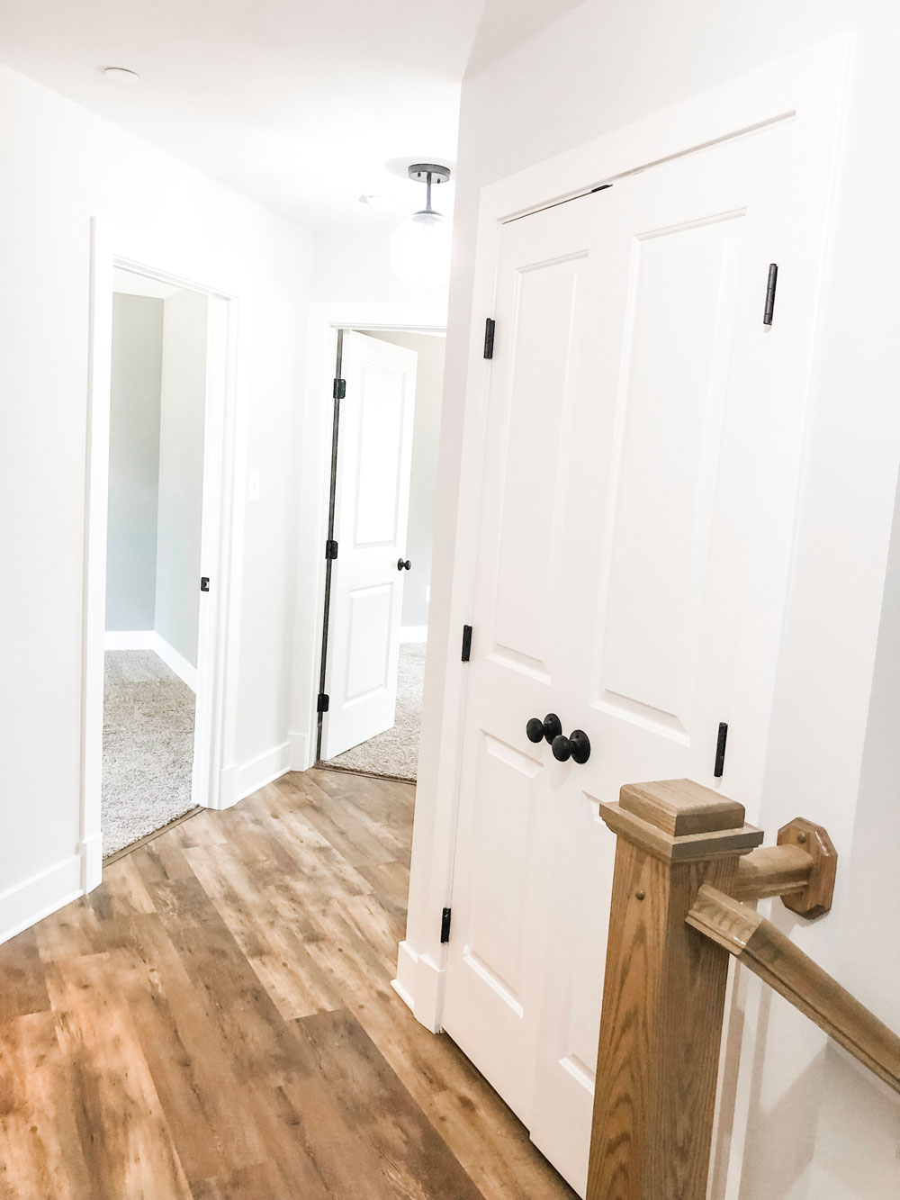 Custom home tour photos. This photo shows the upstairs hallway. The hall has luxury vinyl flooring in a medium tone. There is a large two door linen closet with black knobs.