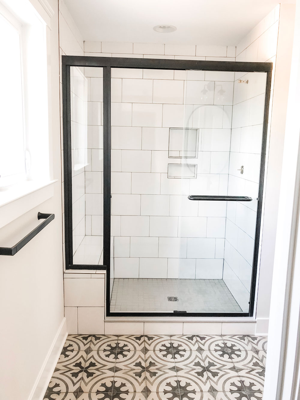 Custom home tour photos. This photo shows the master bath shower. It has a light gray floor tile and large white tiles laid in a subway pattern with gray grout. There is also a black semi-frameless shower door. 