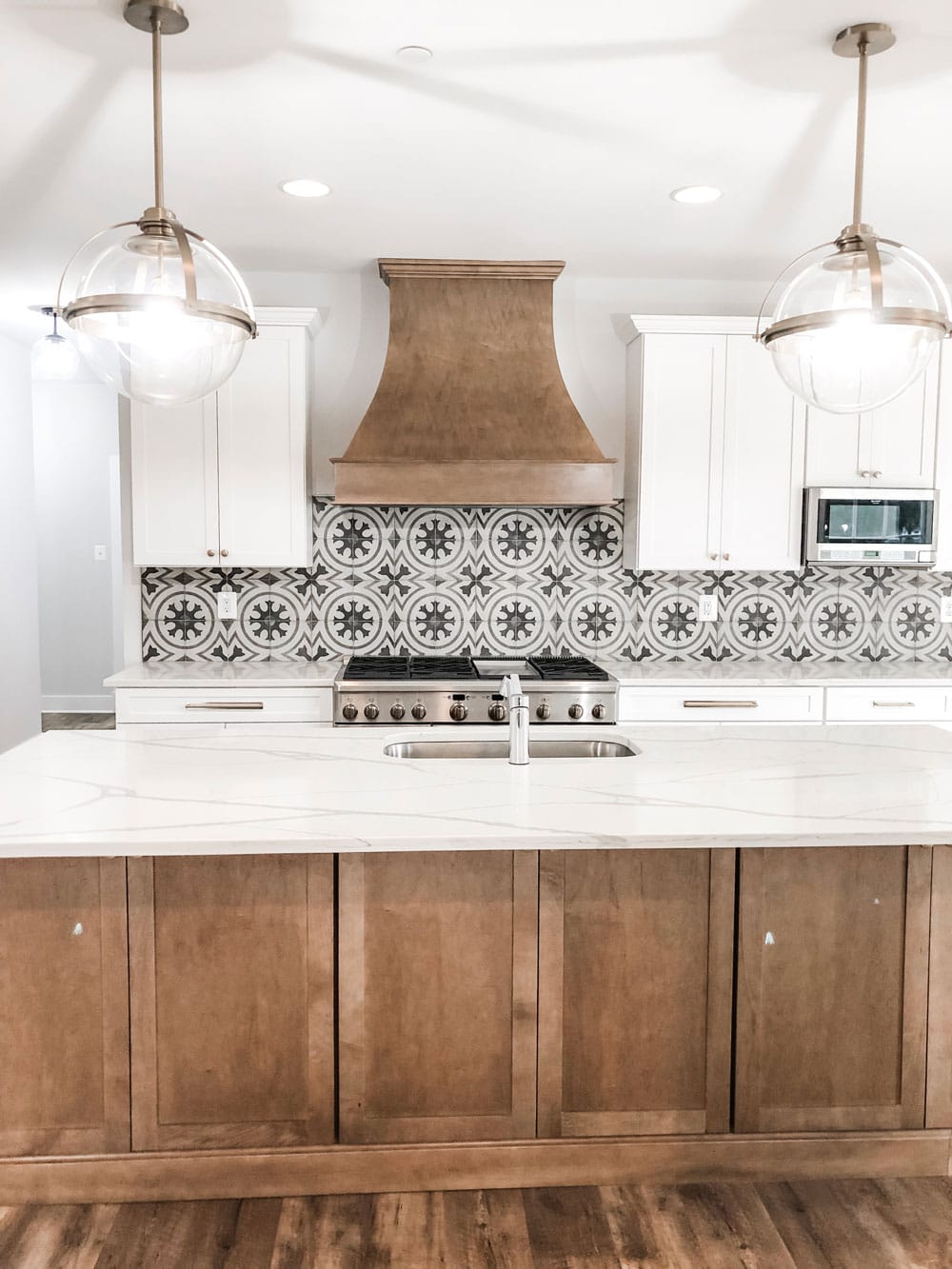 Custom home tour photos. This photo shows the kitchen from the view from the family room. It is looking at the large island with a wood base cabinet and white marble looking counter with gray graining. There are two large glass pendants hanging and a large wood hood above a large 48 inch oven. There are white wall cabinets showing and a stainless microwave. There is also a backsplash with a gray pattern on it. 