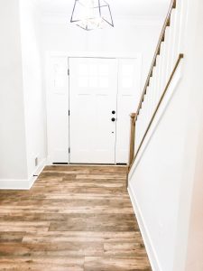 Custom home tour photos. This photo shows the foyer of the home. It has a medium color luxury vinyl wood-looking floor. You can also see the stair rail and the inside of the front door. It is painted white with a black handle.