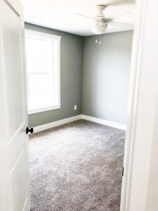 Custom home tour photos. This photo shows one of the two upper bedrooms. It has gray walls and beige carpet. There is a white ceiling fan as well and a large window.