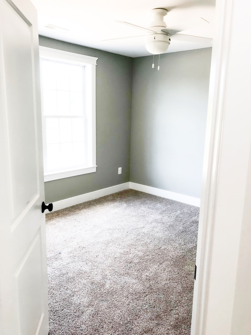 Custom home tour photos. This photo shows one of the two upper bedrooms. It has gray walls and beige carpet. There is a white ceiling fan as well and a large window.