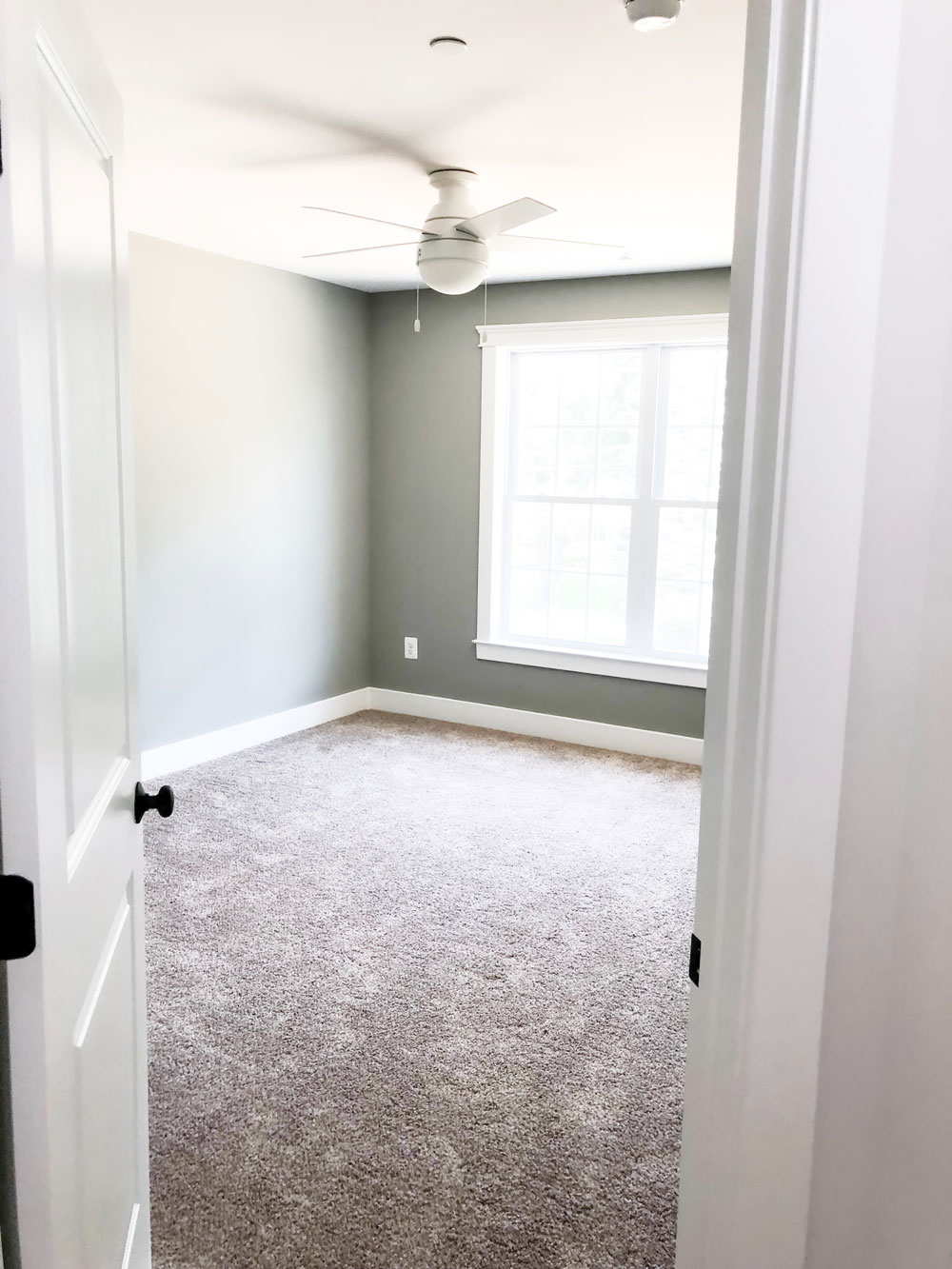 Custom home tour photos. This photo shows one of the two upper bedrooms. It has gray walls and beige carpet. There is a white ceiling fan as well and a large window.