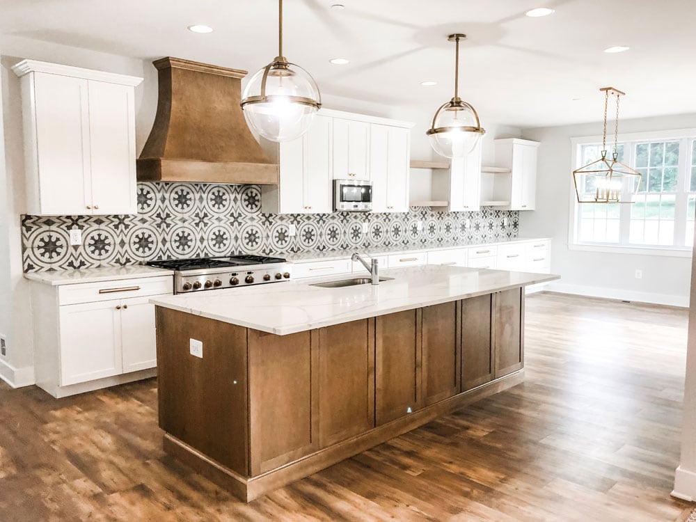 Custom home tour photos. This photo shows the kitchen. It has a large wood island and white cabinets for the rest of the kitchen. There is a large 48 inch range and a wood hood above it. There are two large glass and gold pendants above the island. There is a microwave in the cabinets. There is a gray and white patterned back splash tile. The counters are white with gray graining running throughout them.