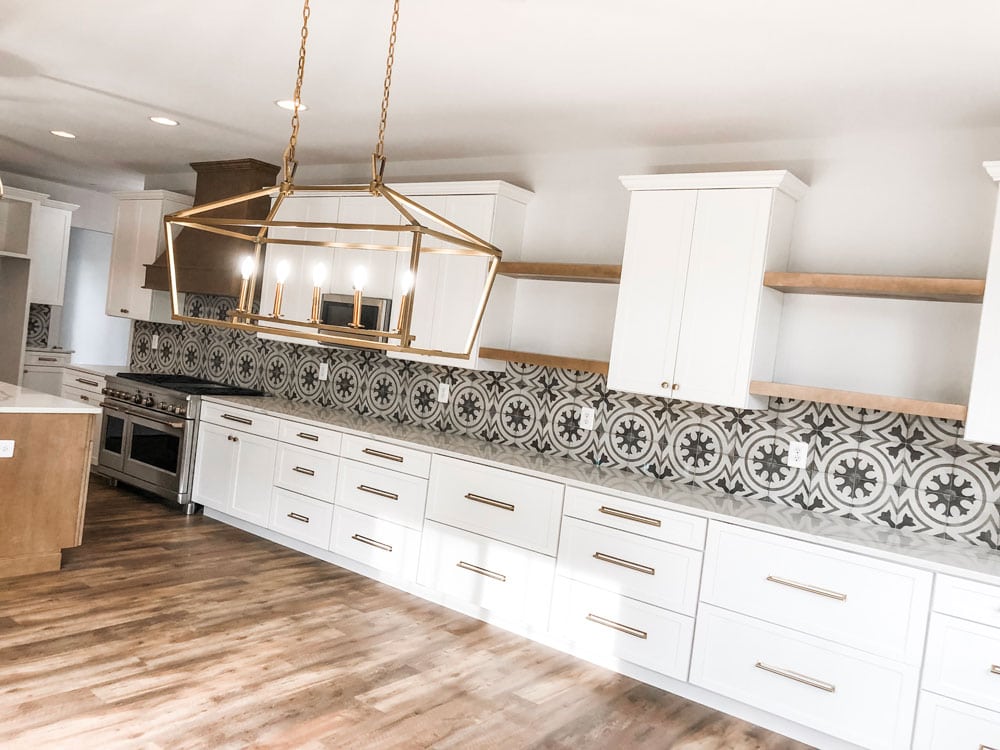 Custom home tour photos. This photo shows a crooked view of the morning room off of the kitchen. There are white cabinets and four wood shelves as well. There is a large gold light that has five lights on it and the light looks like a cage. There are white cabinets with gold pulls and knobs. There is a gray and white patterned back splash.