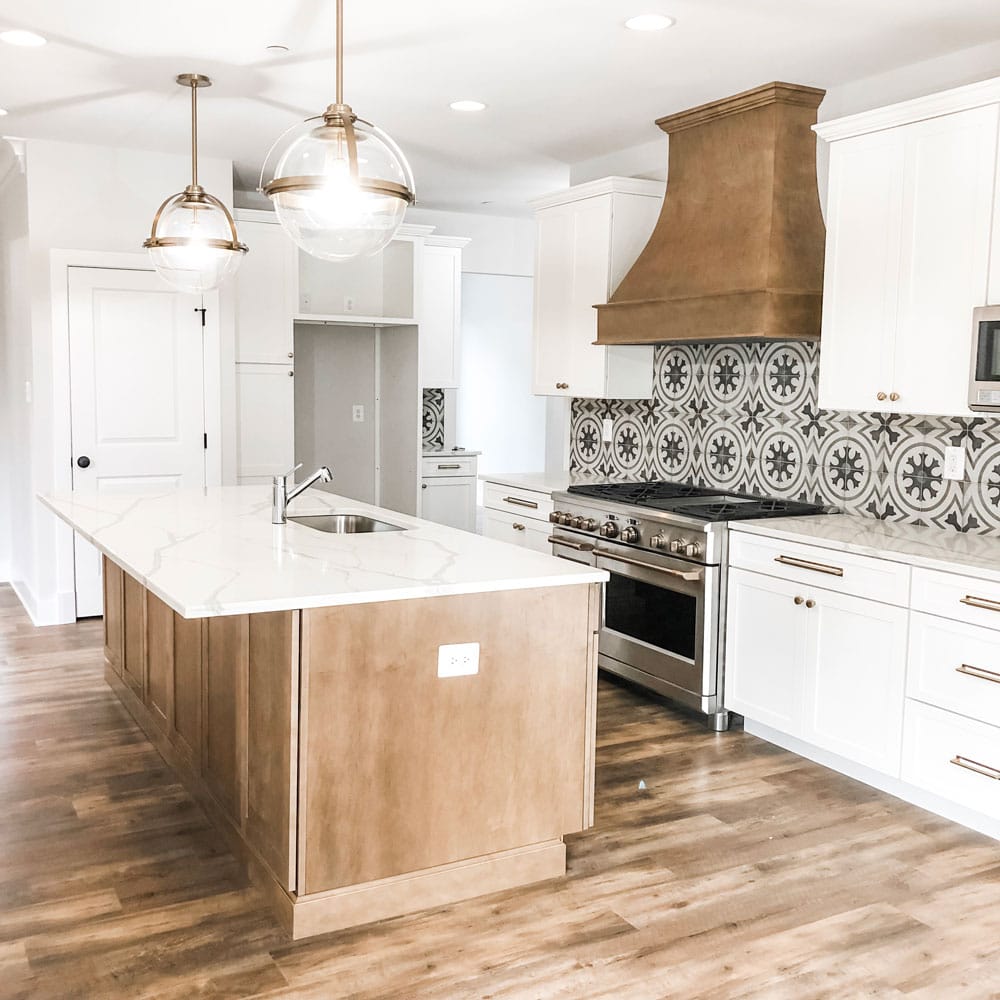 Custom home tour photos. This photo shows the kitchen. It has a large wood island and white cabinets for the rest of the kitchen. There is a large 48 inch range and a wood hood above it. There are two large glass and gold pendants above the island. There is a microwave in the cabinets. There is a gray and white patterned back splash tile. The counters are white with gray graining running throughout them.