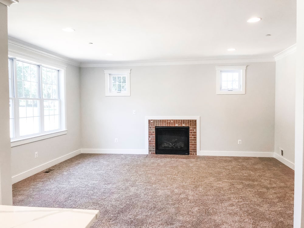 Custom home tour photos. This photo shows the family room. It has white walls, a gas fireplace with red brick surround, beige carpet, two small windows up high on either side of the fireplace, and a window located on the left wall. There is white trim and white crown molding. 