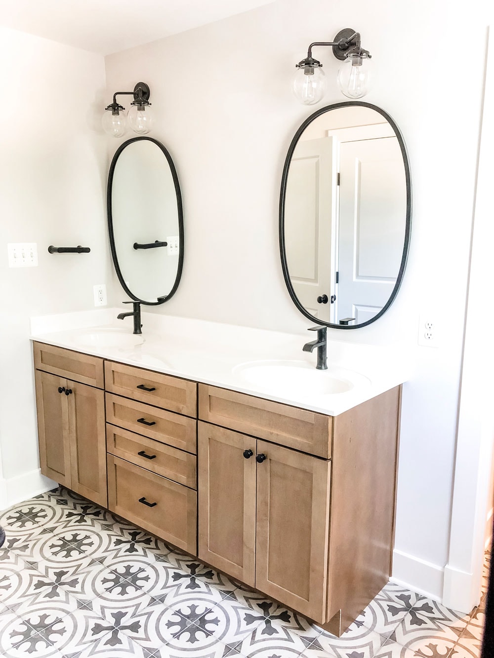 Custom home tour photos. This photo shows the master bathroom with a wood double vanity. The counters are a solid white marble countertop with two sinks. There are two black oval mirrors (one above each sink). There are two black faucets and black hardware on the cabinets. There are also two black lights with glass globes above each mirror. 