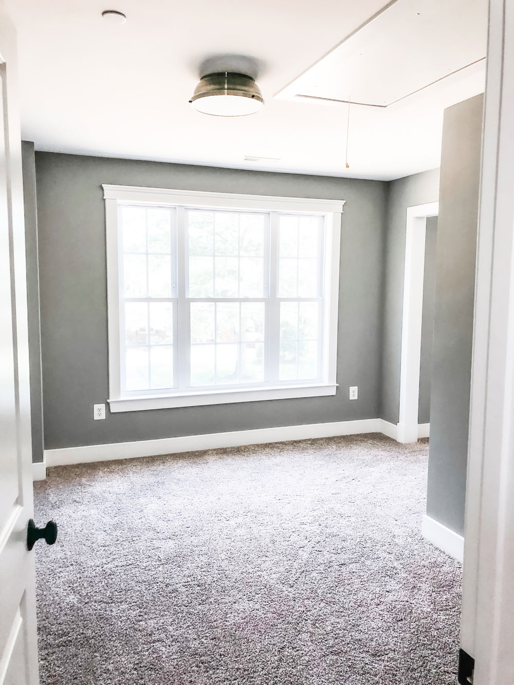 Custom home tour photos. This photo shows the sitting room. It has a gold ceiling light, a large set of three windows, beige carpet, gray walls, and beige carpet. 