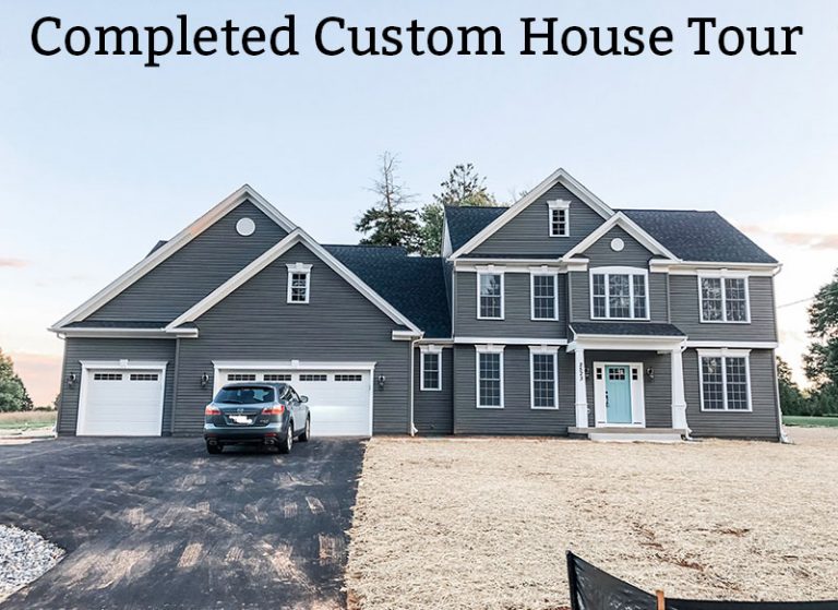 Custom home tour photos. This photo shows the front of the house. The house is a large craftsman with a three-car garage with white garage doors. The house has a blue door and white trim. There is a large driveway with a blue-silver SUV parked in the driveway. There is no grass but instead the yard is full of straw. The house has a small front porch with white tapered columns. The title of the blog post, Completed Custom Tour, across the top center of the picture.