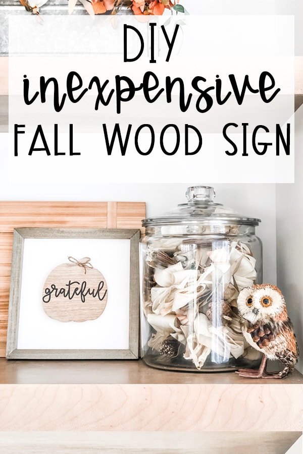 This DIY inexpensive fall wood sign is picture. It's an image of a wood sign with a light wood frame, white background, wood pumpkin, and the words grateful written in black cursive across the pumpkin. A small twine bow is on the stem of the pumpkin. The words, DIY Inexpensive Fall Wood Sign are on the top of the sign. There is also a small fake owl on the far right in front of a glass jar that is filled with dried sticks, leaves, and other outdoor materials in browns and whites.