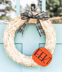 The wreath is on a blue front door, it's made of straw, has a black and white bow at the top, and a bright orange cutout pumpkin with black letters saying, EEK!
