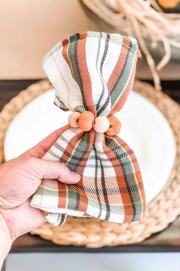 The image is of DIY Thanksgiving napkin rings. They're plaid green, orange, and white napkins with mini pumpkins and wood beads around the napkins.