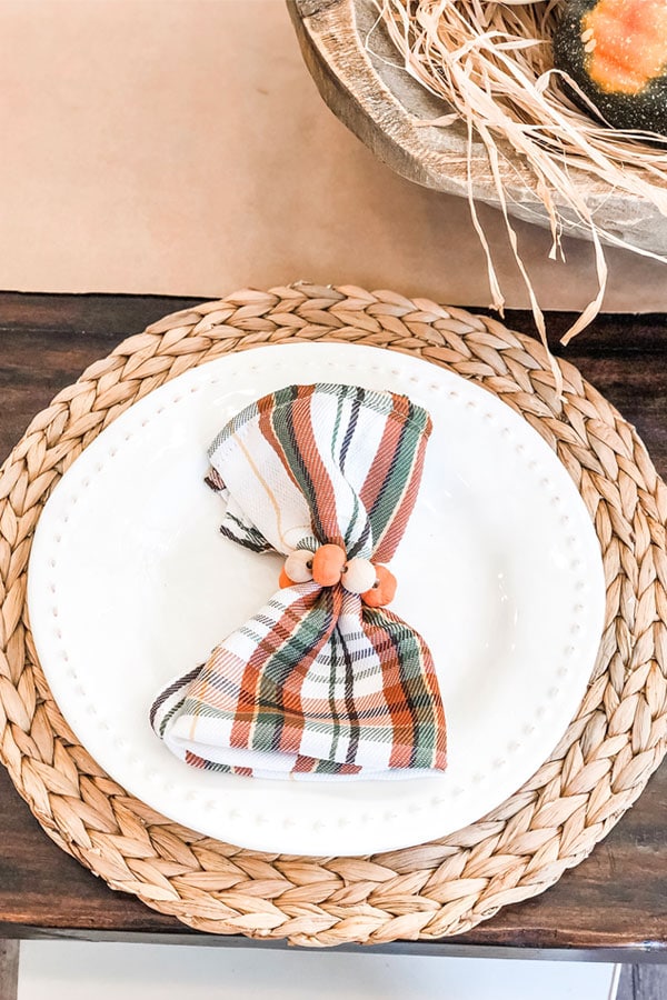 The image is of DIY Thanksgiving napkin rings. They're plaid green, orange, and white napkins with mini pumpkins and wood beads around the napkins. The napkin is sitting on a white plate which is sitting on a straw round placemat, sitting on a dark wood table.