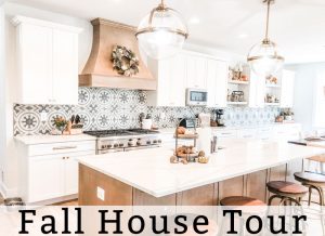 The title fall home tour is at the bottom of the image. Below, is an image of a kitchen. There are two gold and glass pendant lights hanging above an island with a white countertop. The island is a medium wood base. The rest of the cabinets are white. There is a large medium wood hood above the 48 inch stove that is stainless. There is a wreath hanging from the wood hood.