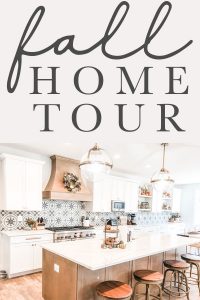 The title fall home tour is at the top of the image. Below, is an image of a kitchen. There are two gold and glass pendant lights hanging above an island with a white countertop. The island is a medium wood base. The rest of the cabinets are white. There is a large medium wood hood above the 48 inch stove that is stainless. There is a wreath hanging from the wood hood.