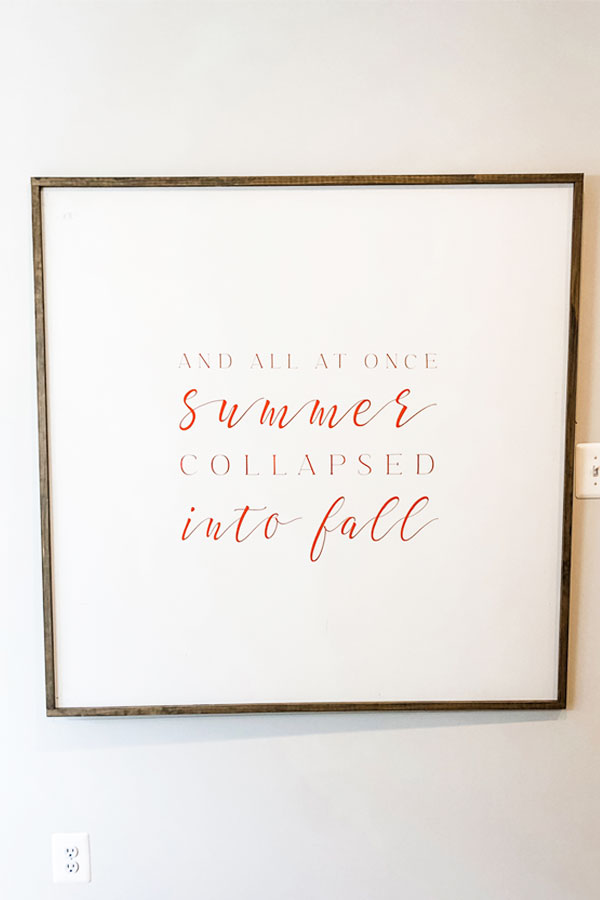 Image of a white wood sign on the wall with the quote, And all at once, summer collapsed into fall, in a mix of orange print and script.