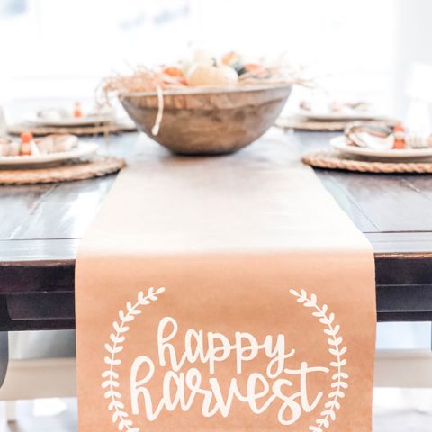 The image shows a kraft paper table runner on a dining table underneath. The words happy harvest are in white on the runner in cursive. There is a small partial wreath on each side of the phrase, happy harvest.