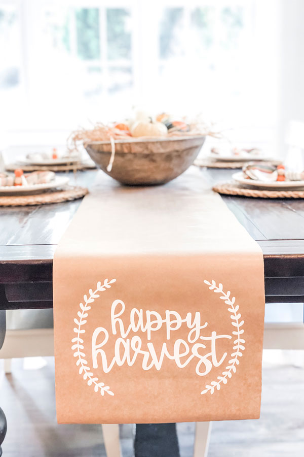 The image shows a kraft paper table runner on a dining table underneath. The words happy harvest are in white on the runner in cursive. There is a small partial wreath on each side of the phrase, happy harvest.