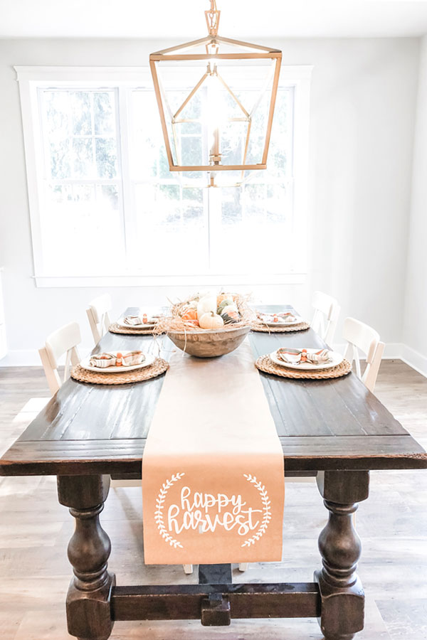 The image shows a kraft paper table runner on a dining table underneath. The words happy harvest are in white on the runner in cursive. There is a small partial wreath on each side of the phrase, happy harvest.