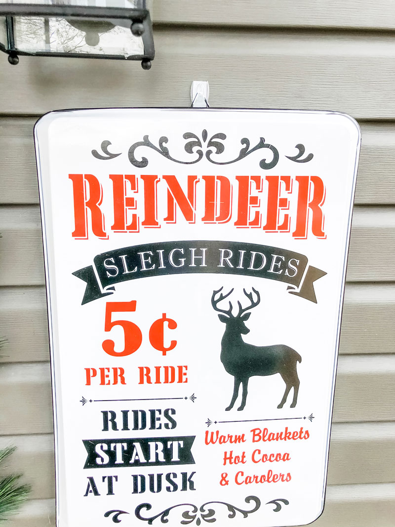 #shop This image shows a sign with the words: Reindeer sleigh rides 5 cents per ride, Rides start at dusk. Warm blankets, hot cocoa, & carolers with gray siding behind it.