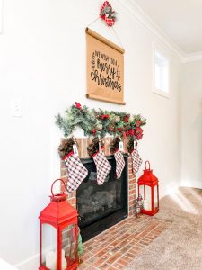 #shop This image shows a brick fireplace surround with two large red lanterns sitting on the ground on either side. Four stockings are hung above the fireplace with a garland above that. Above the garland is a sign with black letters on kraft paper saying: we wish you a Merry Christmas. There is a swag of greenery and a red and white checkered bow. Next to the red lanterns on the floor are a green and red plaid reindeer, green tree, and two plaid trees. Each stocking has a name hanger in white that say: Dad, Mommy, Carl, Jack.