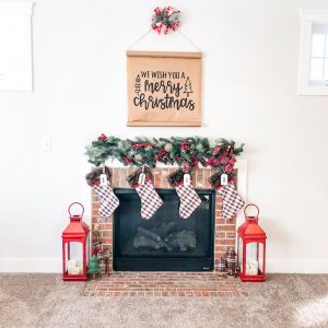#shop This image shows a brick fireplace surround with two large red lanterns sitting on the ground on either side. Four stockings are hung above the fireplace with a garland above that. Above the garland is a sign with black letters on kraft paper saying: we wish you a Merry Christmas. There is a swag of greenery and a red and white checkered bow. Next to the red lanterns on the floor are a green and red plaid reindeer, green tree, and two plaid trees. Each stocking has a name hanger in white that say: Dad, Mommy, Carl, Jack.