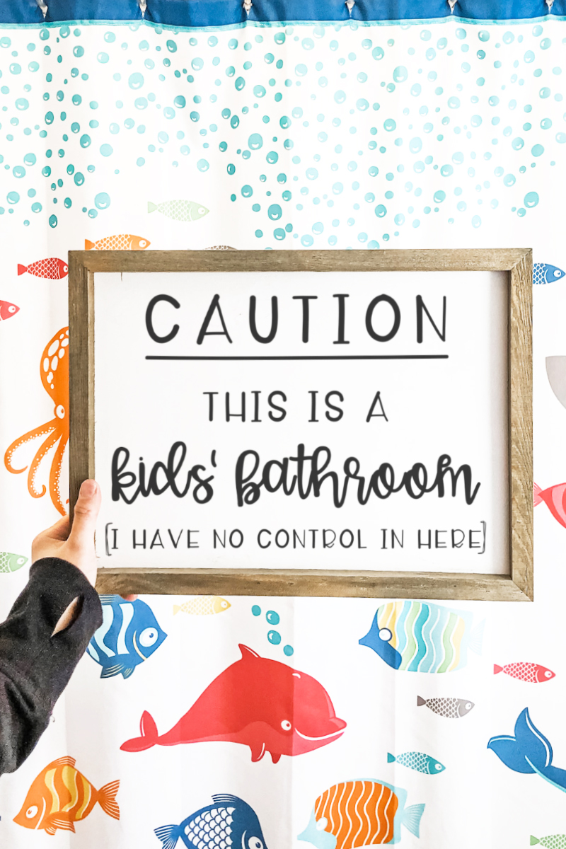 Funny Bathroom SVG and Print at the top in black text with a white background. Below it is a white sign with a wood frame. The sign in black text reads: Caution this is a kids' bathroom (I have no control in here). The background behind the sign is a white shower curtain with brightly colored fish and blue bubbles.