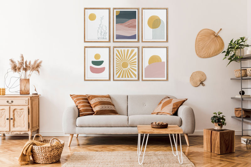 Living room with a beige couch, brown pillows, and a small wood and white coffee table in front of the couch. There is a small beige rug in the center of the room. To the left of the couch is a small sideboard with a feather flower decor. To the right of the couch are two fans on the wall. To the right of that is a black pipe and wood shelf bookshelf with various pieces of decor. On the floor to the right of the coffee table is a small wooden cube table with a plant on it. To the left of the couch is a basket with a blanket within in. Above the couch are 6 abstract, boho prints in colors of pinks, yellow, and green.