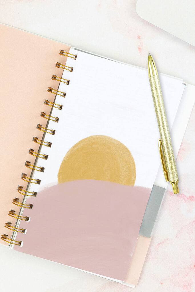 Open pink planner with an abstract divider showing a yellow sun with a large pink cloud-like shape overtop of it. With a gold pen on top of the planner.