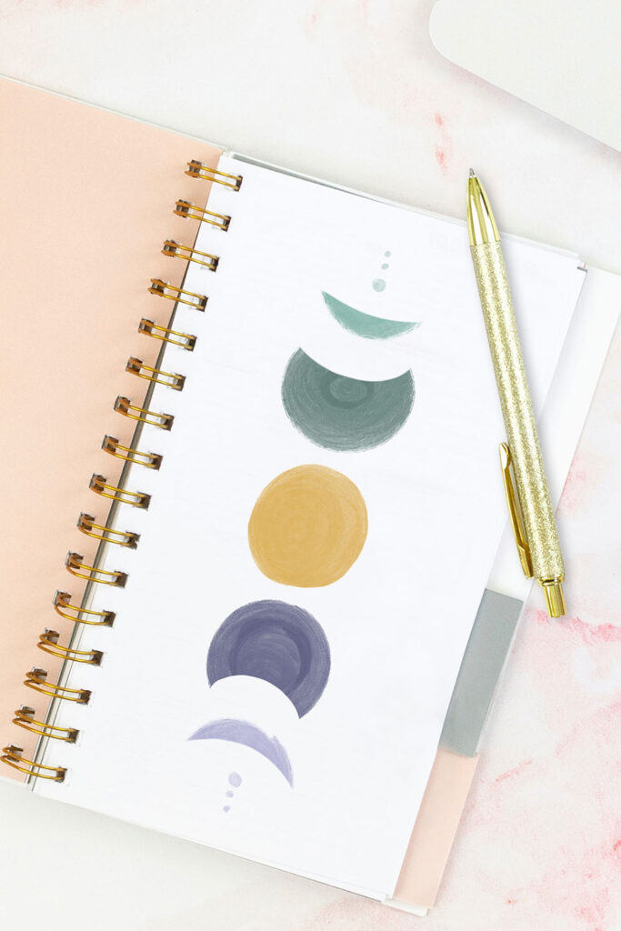 Open pink planner with an abstract divider showing a yellow moon in the center with two 3/4 moons on either side in purple and green, and then two crescent moons on either side in purple and green, and two tiny circles in green and purple on either side of that . With a gold pen on top of the planner.