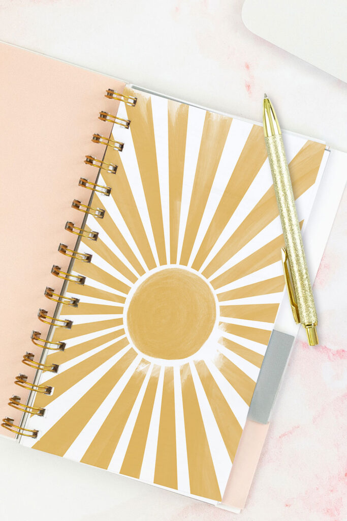 Open pink planner with an abstract divider showing a yellow sunburst. With a gold pen on top of the planner.