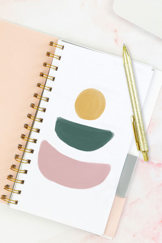 Open pink planner with an abstract divider showing a yellow sun over top of an abstract green shape similar to a bowl or half-circle, and a pink abstract shape also shaped like a bowl or half-circle. With a gold pen on top of the planner.