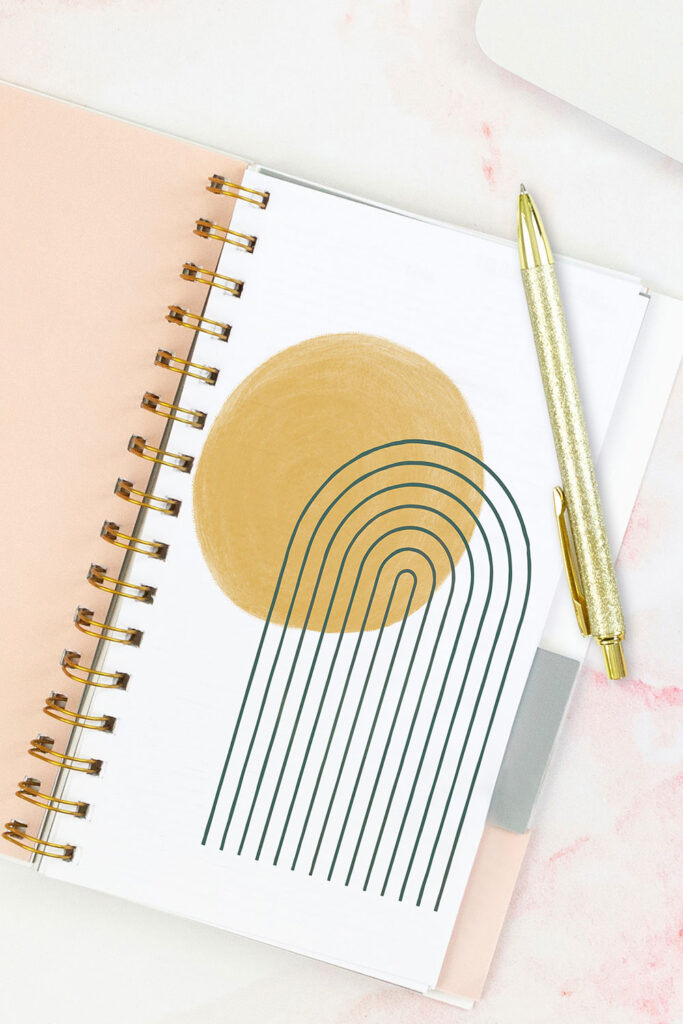 Open pink planner with an abstract divider showing a yellow sun and a dark blue rainbow. With a gold pen on top of the planner.