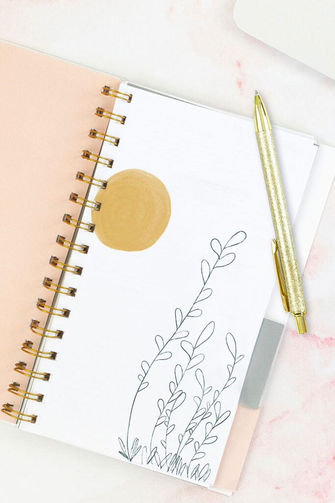 Open pink planner with an abstract divider showing a yellow sun and green plant. With a gold pen on top of the planner.