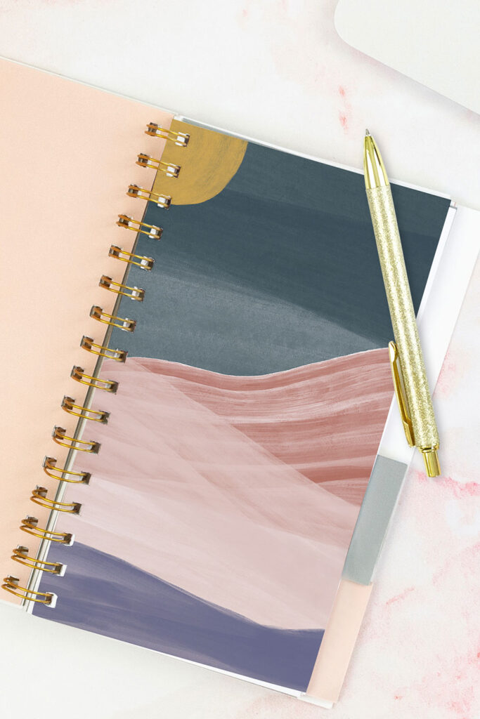 Open pink planner with an abstract divider showing a yellow moon, dark blue sky, and pink/purple mountains. With a gold pen on top of the planner.