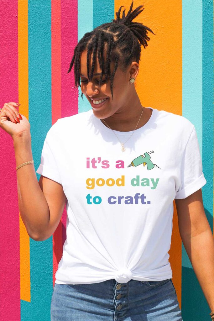 Woman standing in a white t-shirt and jeans smiling and looking off to the side. She has one arm down to her side and another raised. She's up against a bright, colorful striped background. Her t-shirt says it's a good day to craft with a glue gun.