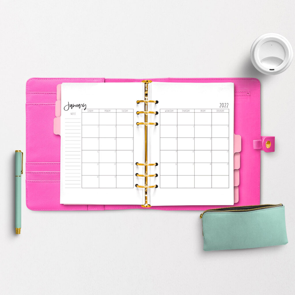 The image shows the 2022 Free Printable Calendar you can download. The image is of an open planner with a bright pink cover. Inside of the planner are pink tab dividers and it's open to a two page January 2022 calendar. On the top right is the lid of a white travel mug. To the bottom left next to the open planner is a mint green and gold pen. Below the planner to the bottom right is a mint green pencil pouch with a gold zipper. The pouch is zipped open. 