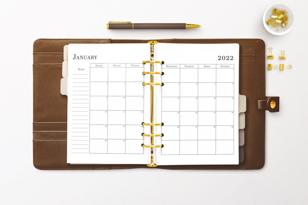 A brown leather planner lays open. Inside is a January 2022 calendar spread in black text on white paper. There are gold rings in an A5 style. A brown pen sits just above the planner. To the right are gold binder clips in a small bowl and four lay on the table below the bowl.