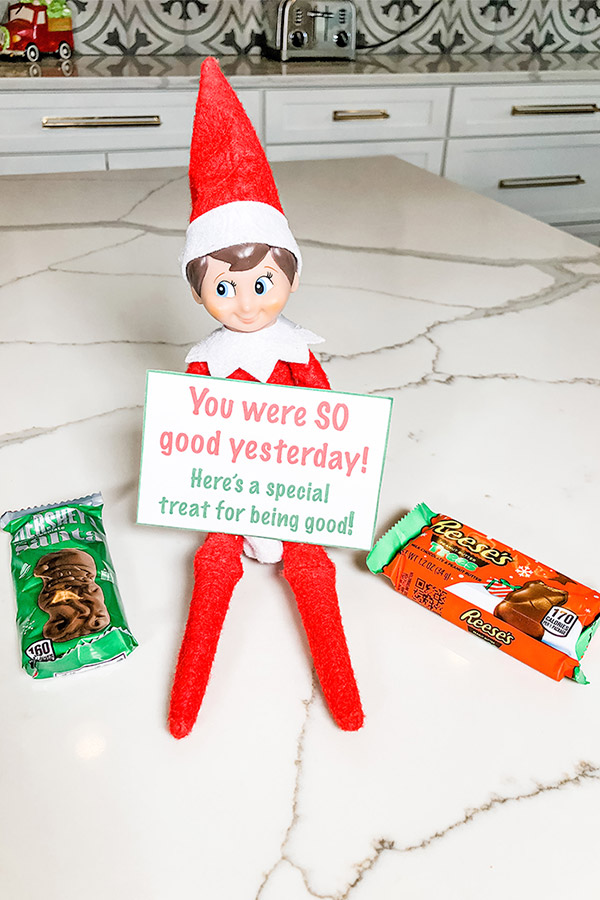 An elf on a shelf doll is sitting holding a note that says, "you were SO good yesterday! Here's a special treat for being good!" Next to the elf is a small Hershey bar shaped like Santa in it's wrapper, and a Reese's Christmas tree in it's packaging. 