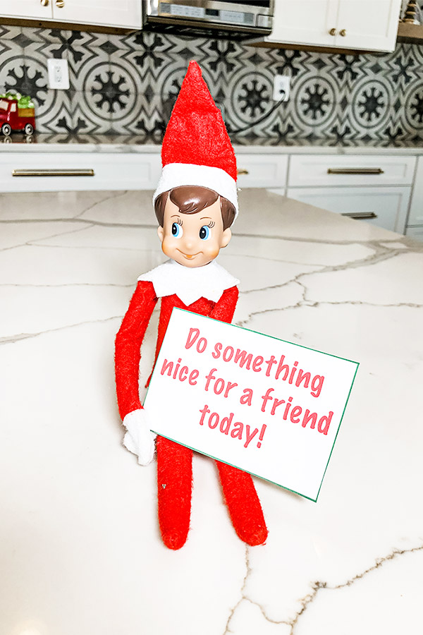 An elf on a shelf doll is sitting holding a note that says, "do something nice for a friend today!"
