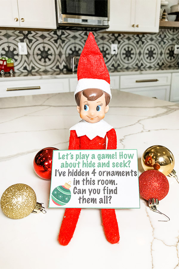 An elf on a shelf doll is holding a note that says, "let's play a game! How about hide and seek? I've hidden 4 ornaments in this room. Can you find them all?" Next to the elf are some red and gold glitter ornaments. 