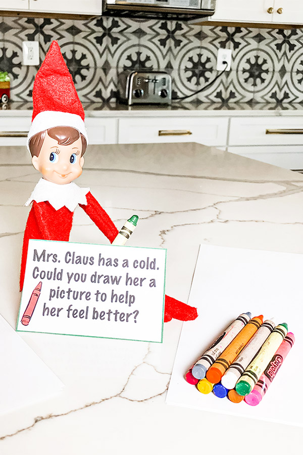 An elf on the shelf doll is sitting, holding a green crayon and a note that says, "Mrs. Claus has a cold. Could you draw her a picture to help her feel better?" Next to the elf is a stack of crayons on some white printer paper.