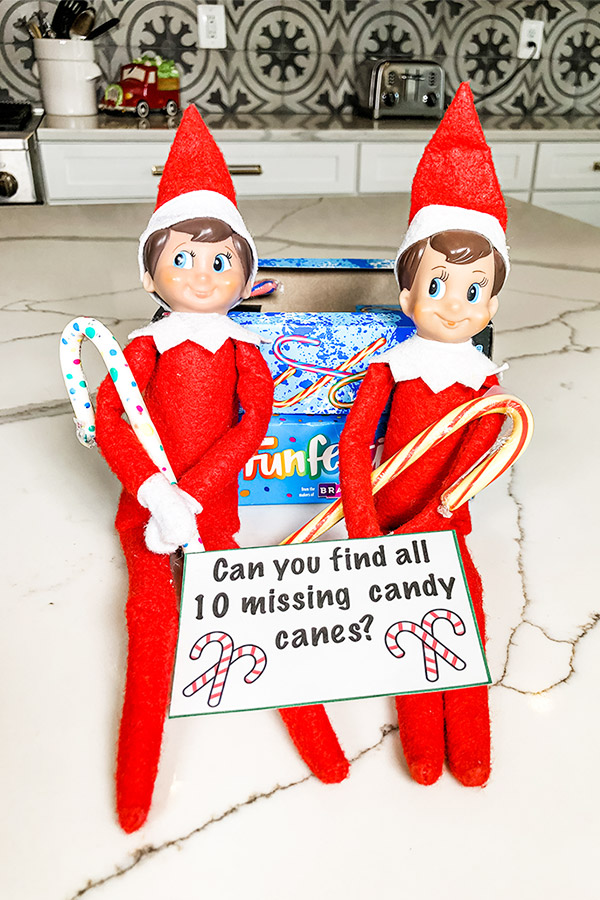 Two elf on the shelf dolls are sitting holding candy canes. They have a note with them that says, "Can you find all 10 missing candy canes?" They're sitting against two candy cane boxes.