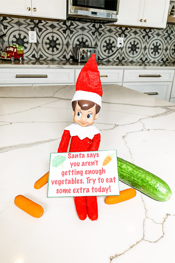 An elf on the shelf doll is sitting on a counter with a note that says, "Santa says you aren't getting enough vegetables. Try to eat some extra today!" Next to the elf are 3 baby carrots and a small green cucumber.