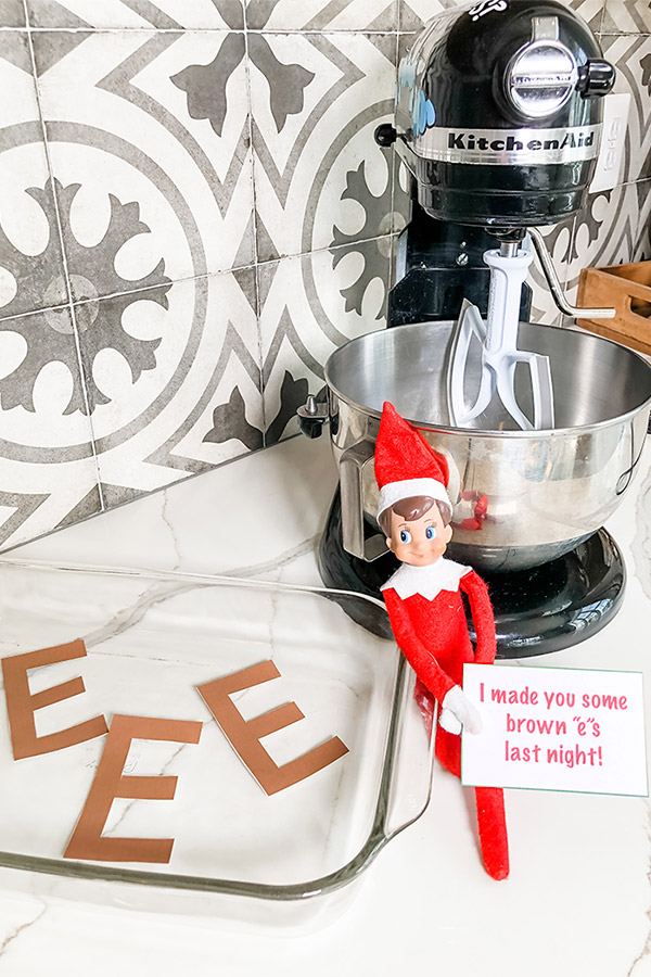 An elf on the shelf doll is sitting against a kitchenaid stand mixer holding a note that says "I made you some brown "e"s last night!" Next to the elf is a clear pyrex baking dish with brown paper letter e's. They're cut out and laying in the dish to represent brown "e"s as a joke like they're brownies.